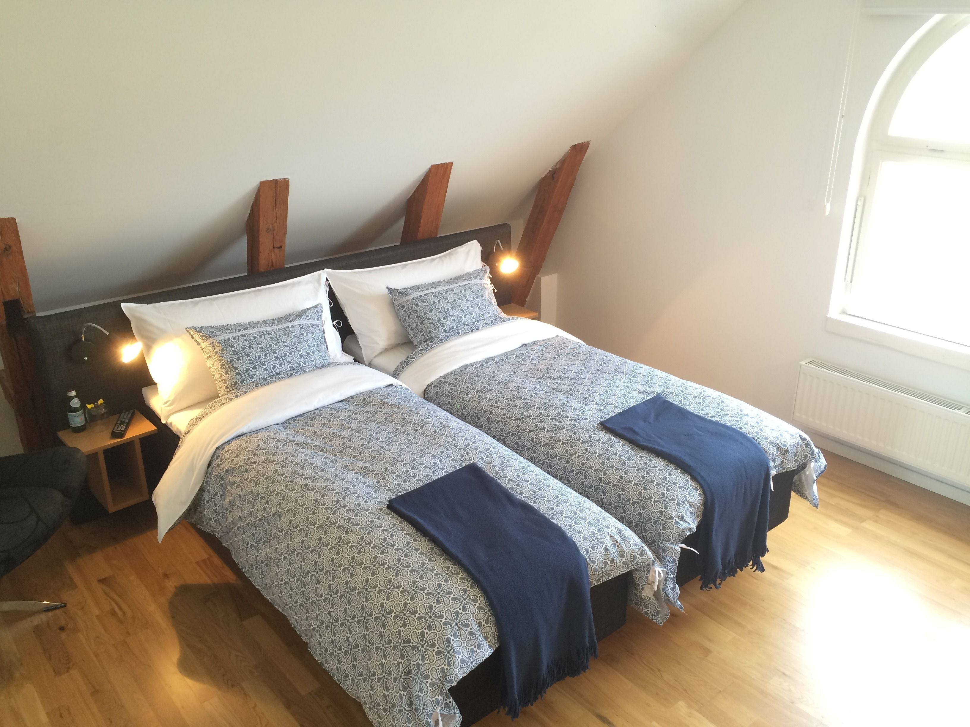 A bedroom with two beds, wooden headboard, bedside lamps, and a window with natural light.