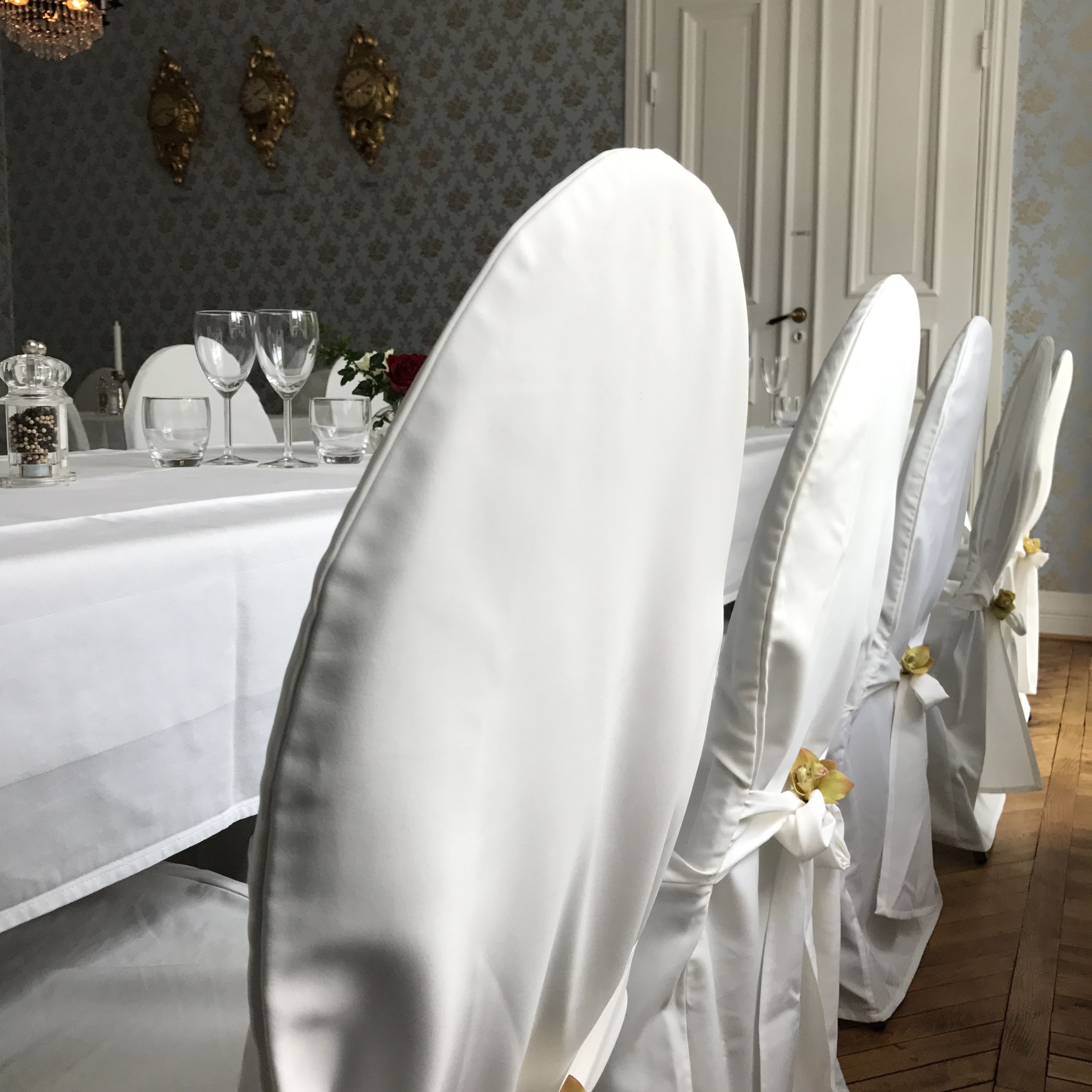 A dining table with white tablecloth and glassware, set with white chairs adorned with yellow flowers.