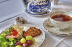 A plate with cheese, ham, lettuce, and grapes, a heart-shaped cookie, and a cup of tea on a table with a blue and white teapot in the background. - Annas Hotell (Kristianstad)