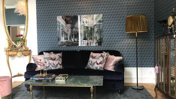A living room with a dark blue patterned wall, a black velvet sofa with floral cushions, a glass coffee table, a gold-framed mirror, and a large framed picture above the sofa.