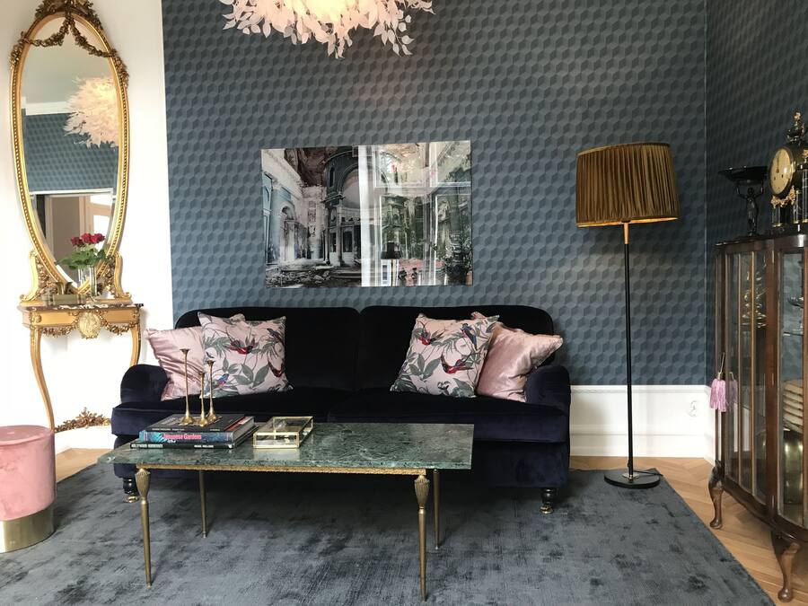 En stue med en mørkeblå væg med mønster, en sort veloursofa med blomstermønstrede puder, et sofabord i glas, et spejl med gylden ramme og et stort billede hængende over sofaen.