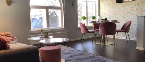A modern living room with a round table, two pink chairs, a sofa, and a large window with a view of the outside.