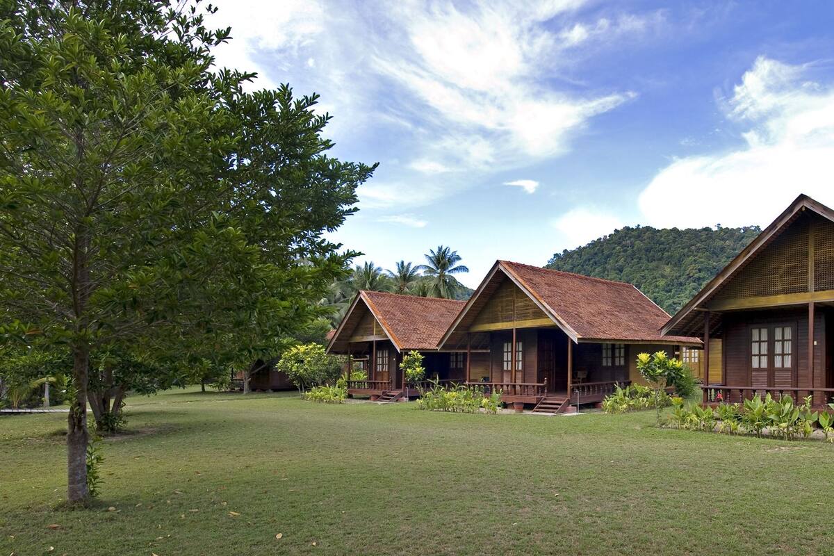 Une rangée de cabanes en bois aux toits en pente, entourées d’une végétation luxuriante et de palmiers, se dressant sur fond de paysage montagneux.