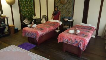 A room with two beds, a small table, a chair, and a decorative wall.