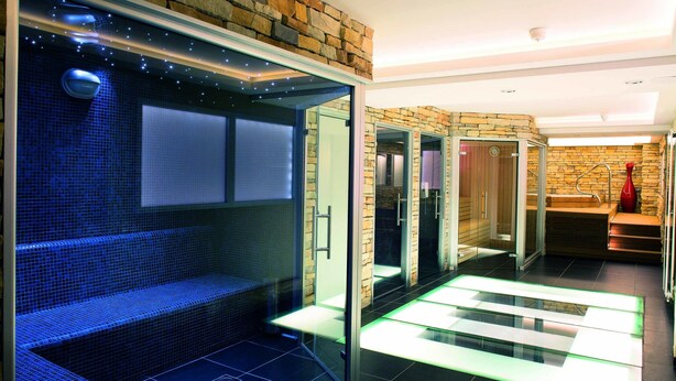 A modern spa area with a glass-enclosed sauna, a blue-tiled bench, and a glass floor leading to a shower area.