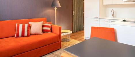 A modern living room with an orange sofa, a small round table, and a kitchen area with white cabinets.