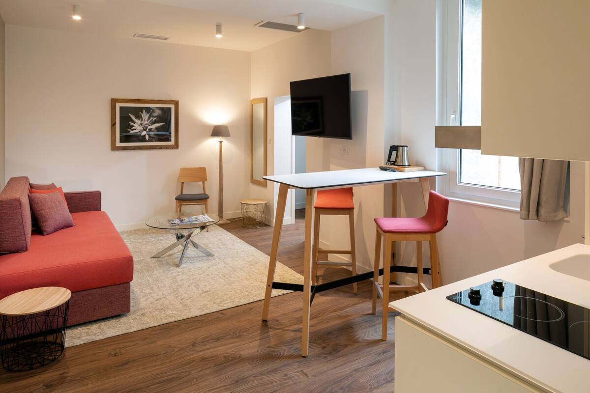 A modern living room with a red sofa, a glass coffee table, a wooden dining table with two chairs, a TV mounted on the wall, and a kitchen area with a stove.