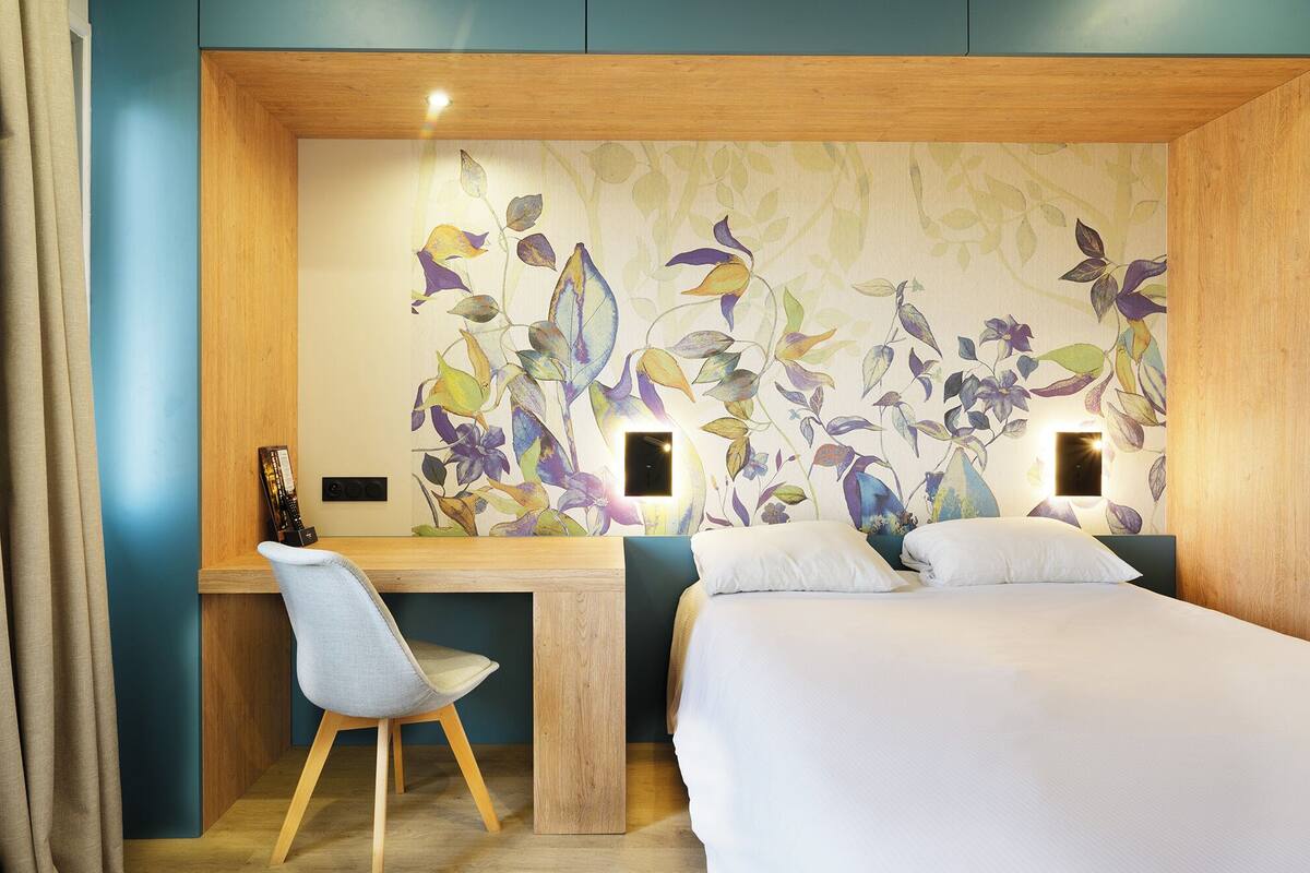 A hotel room with a wooden desk, a chair, a bed, and a floral wallpaper.