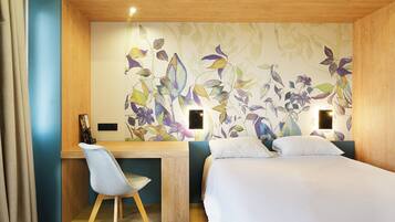 A hotel room with a wooden desk, a chair, a bed, and a floral wallpaper.