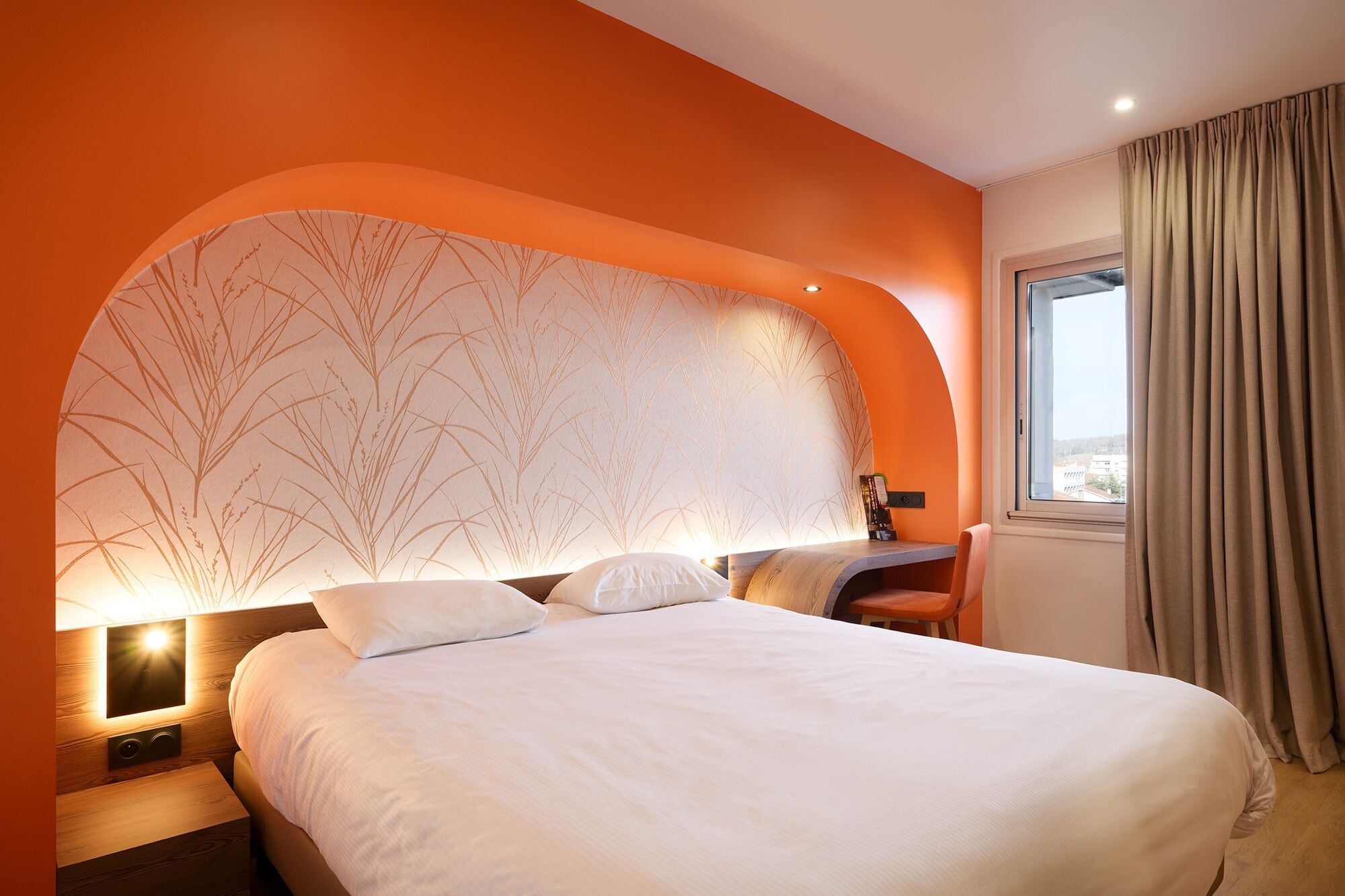 A hotel room with a large bed, a curved headboard with a tree pattern, a nightstand, a chair, and a window with curtains.