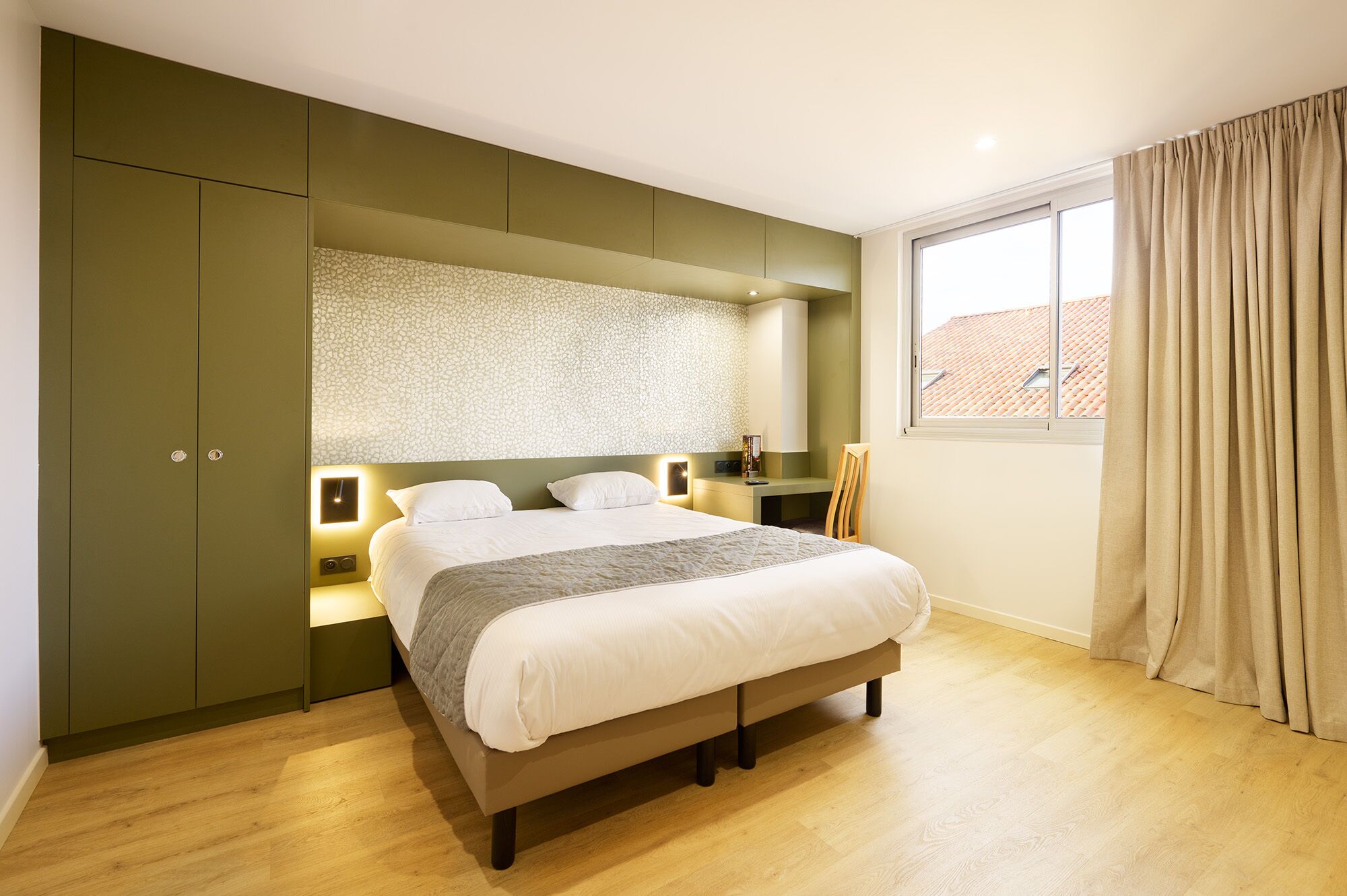 A bedroom with a large bed, a wardrobe, a nightstand, a lamp, and a window with curtains.