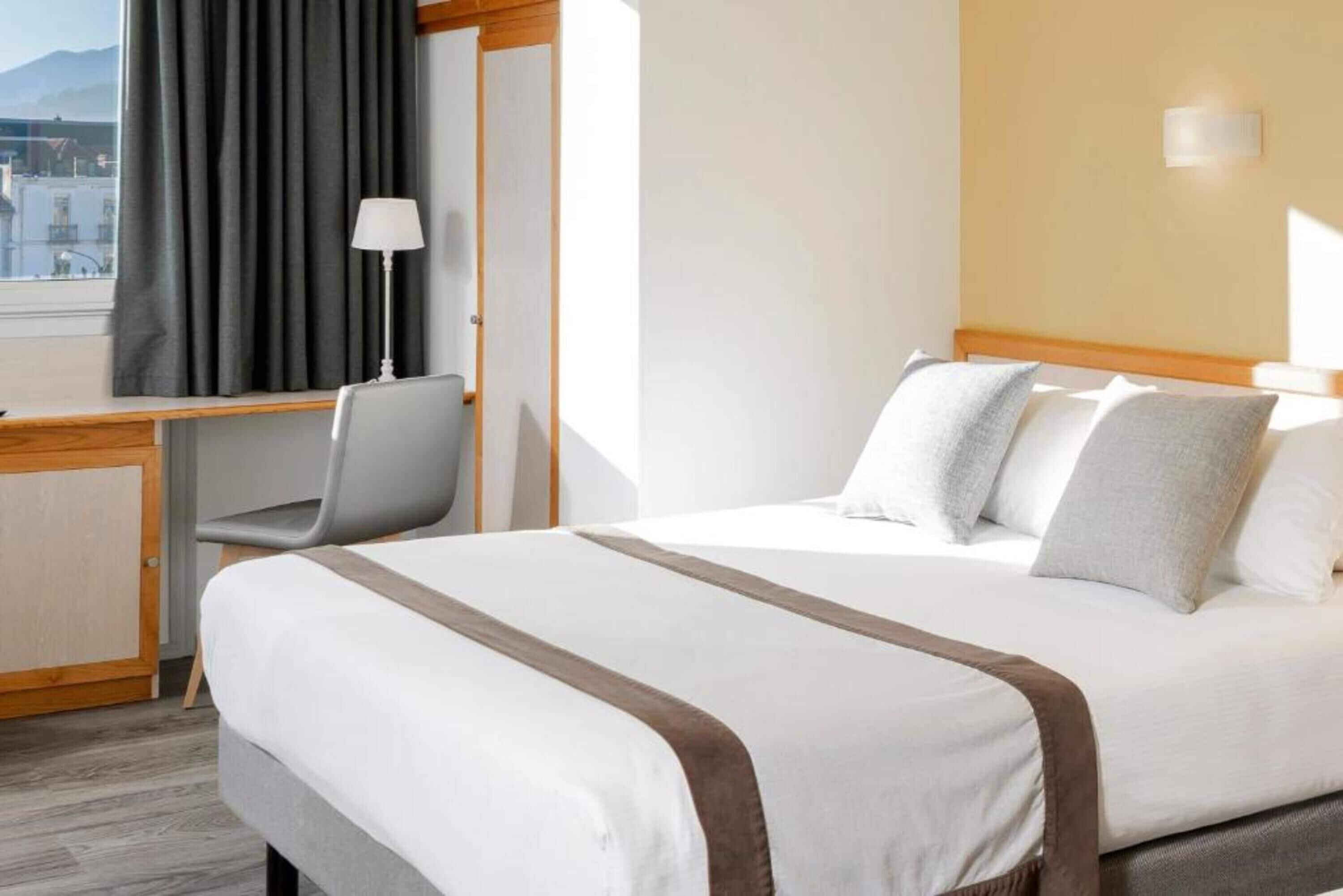 Ett hotellrum med en säng, ett skrivbord, en stol, en lampa och utsikt över staden.