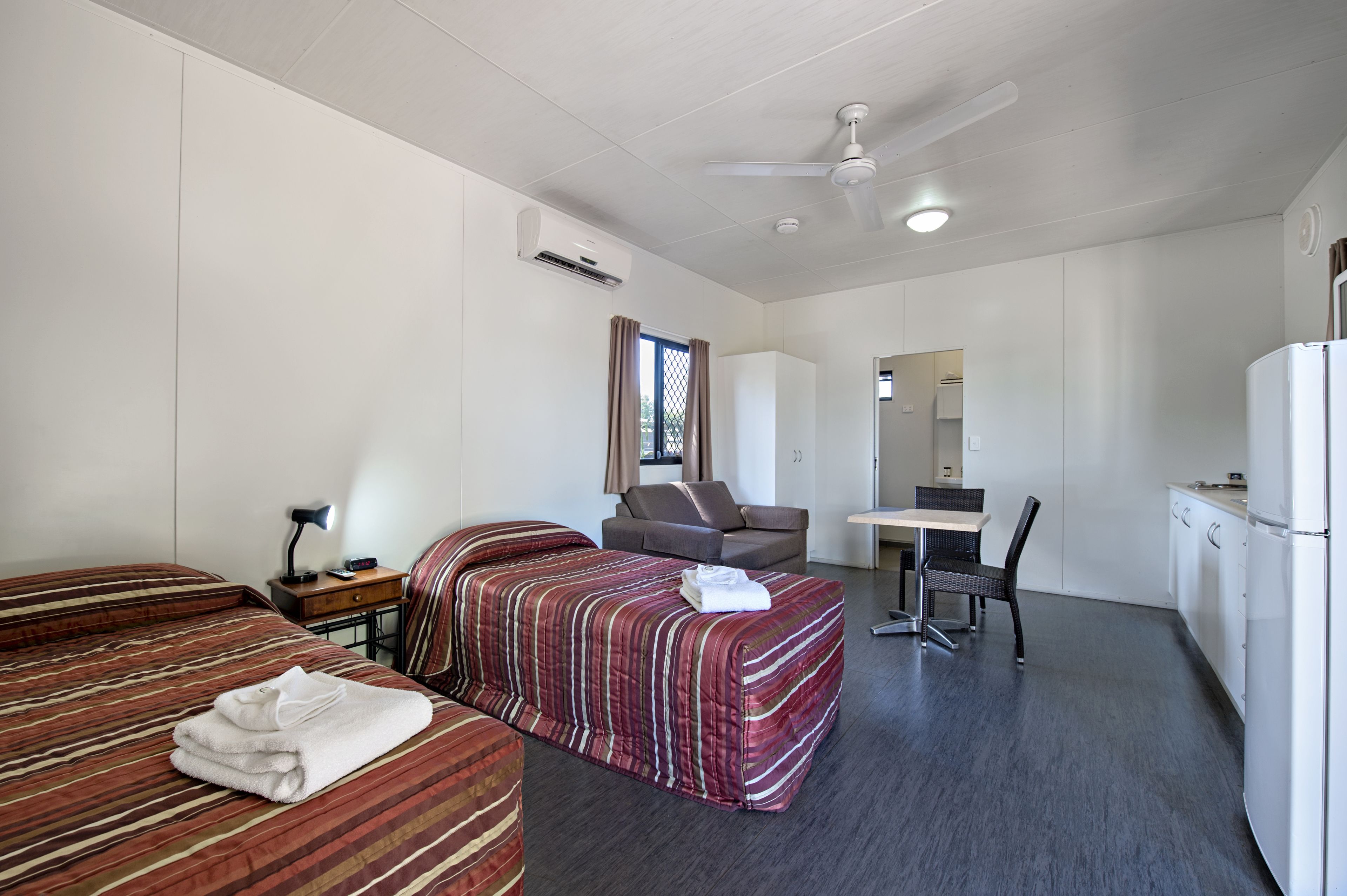 A hotel room with two beds, a desk, a chair, a refrigerator, and a ceiling fan.
