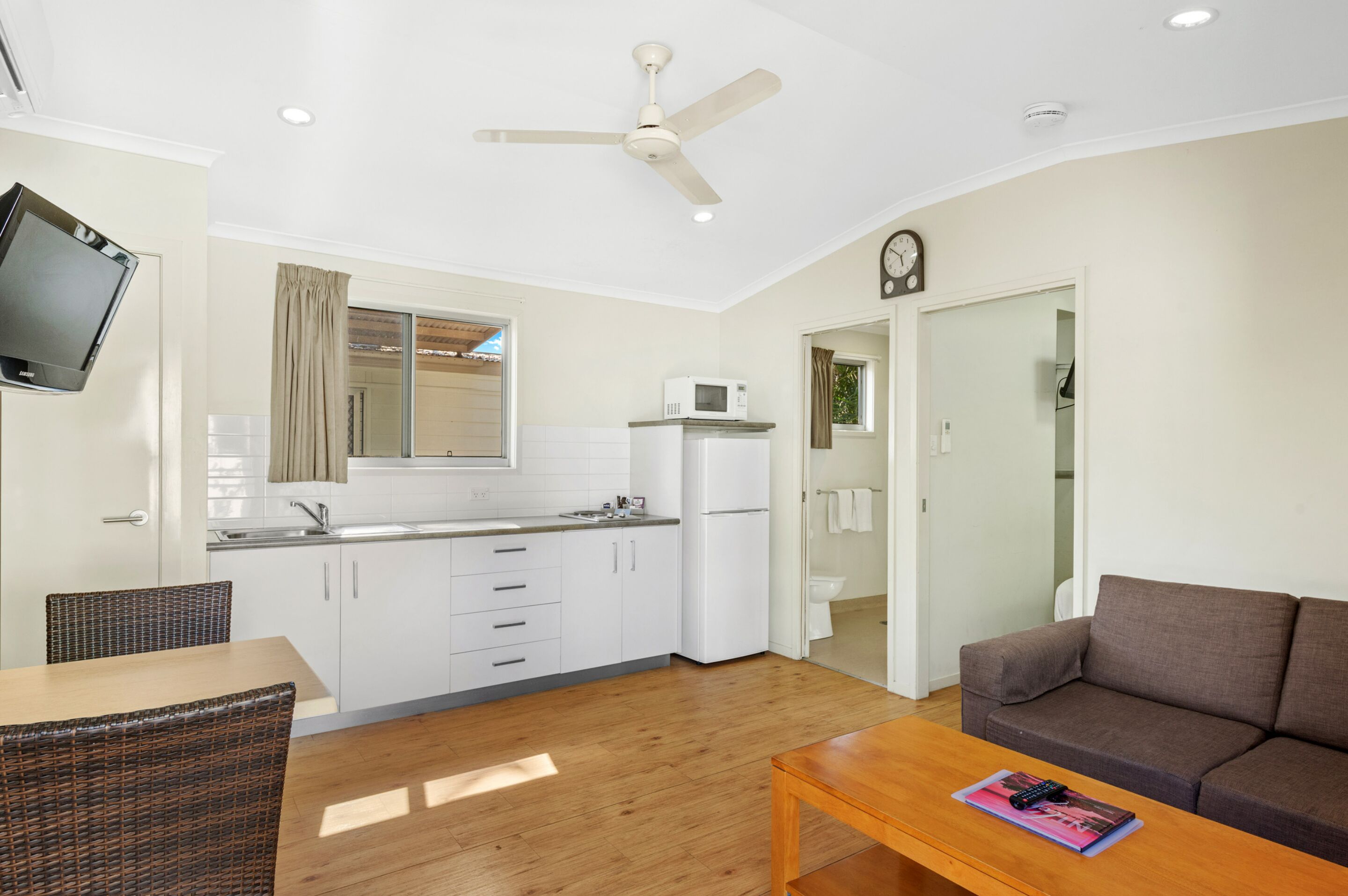 A compact living space with a kitchenette, a dining area with wicker furniture, a sofa, and a ceiling fan.