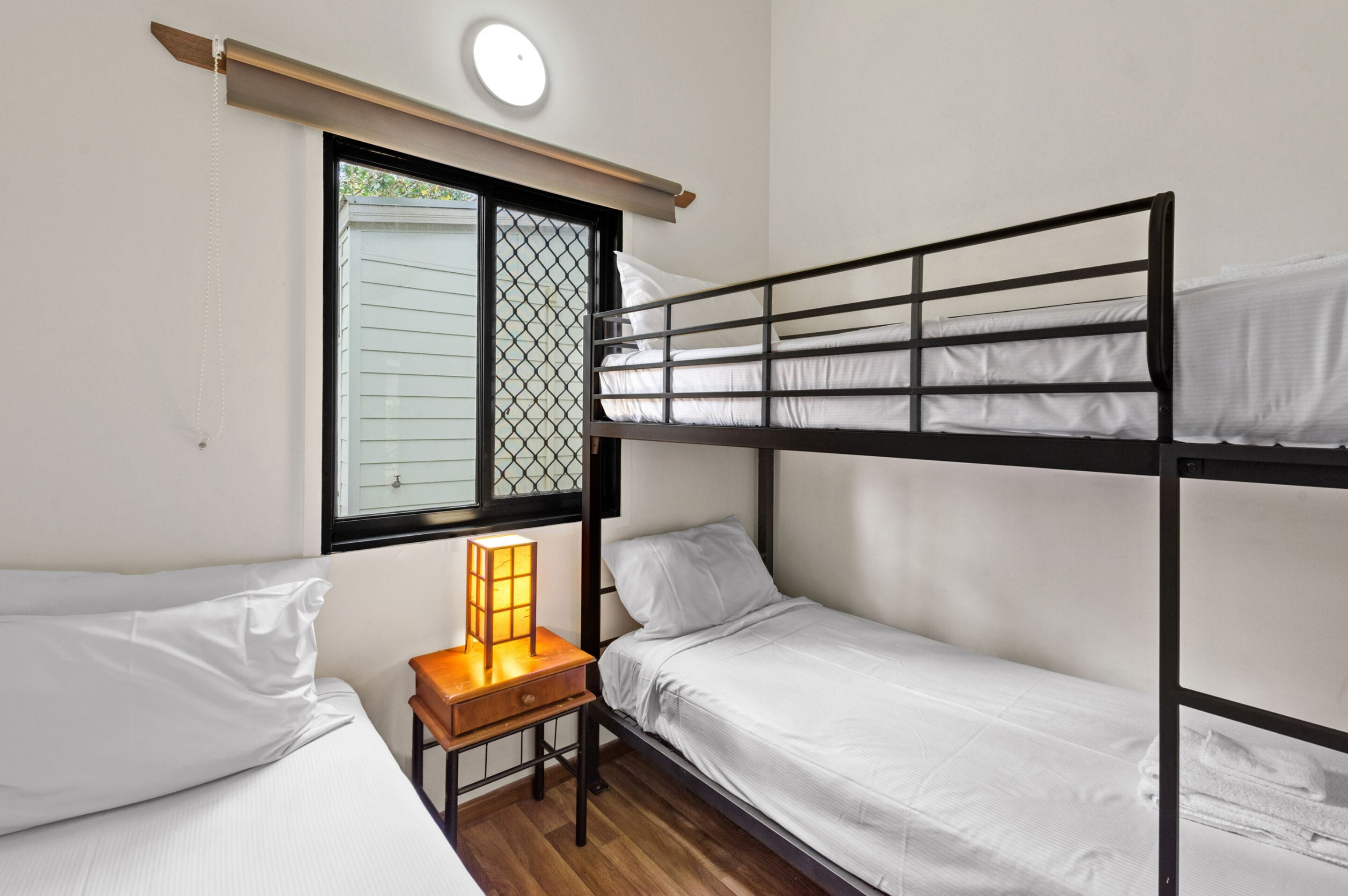 A bunk bed room with a small table and a lamp, a window with blinds, and a ceiling light.
