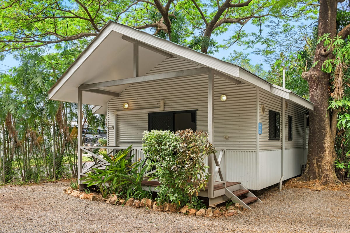 A small, single-story mobile home with a porch, surrounded by trees and gravel.
