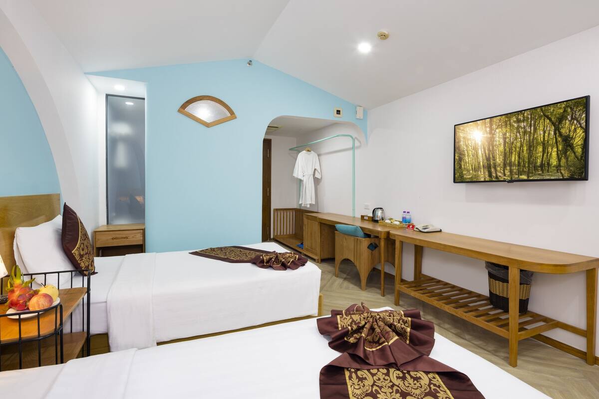 A hotel room with two beds, a desk, a chair, a small table with fruit, a robe, and a painting on the wall.
