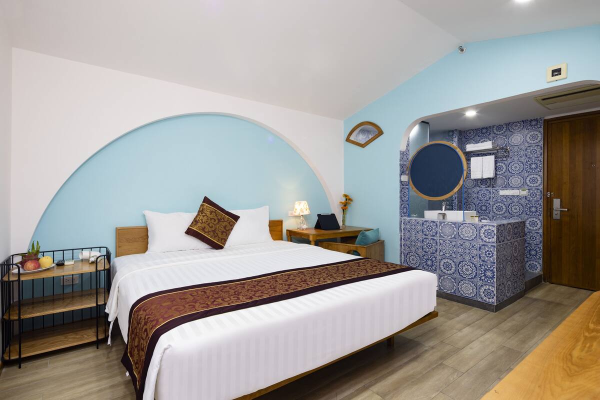 A hotel room with a large bed, a bedside table, a desk, a chair, a small table with fruit, a mirror, a sink, and a door.