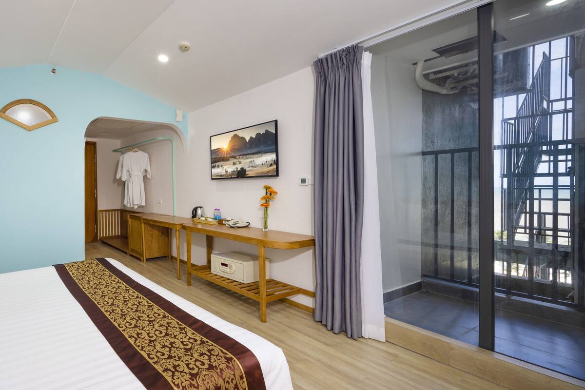 A hotel room with a bed, a desk, a chair, a wardrobe, a window with curtains, and a view of a building through the window.