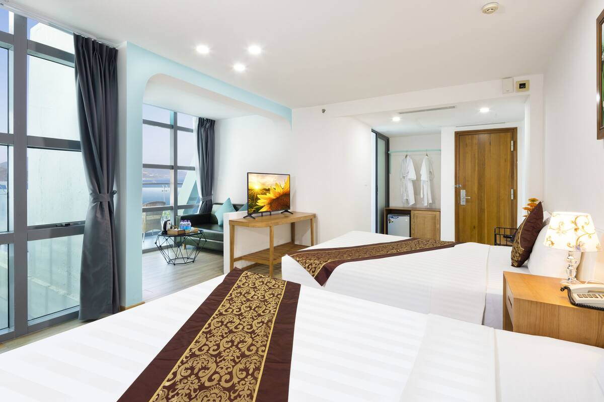 A hotel room with two beds, a TV, a desk, and a view of the sea.