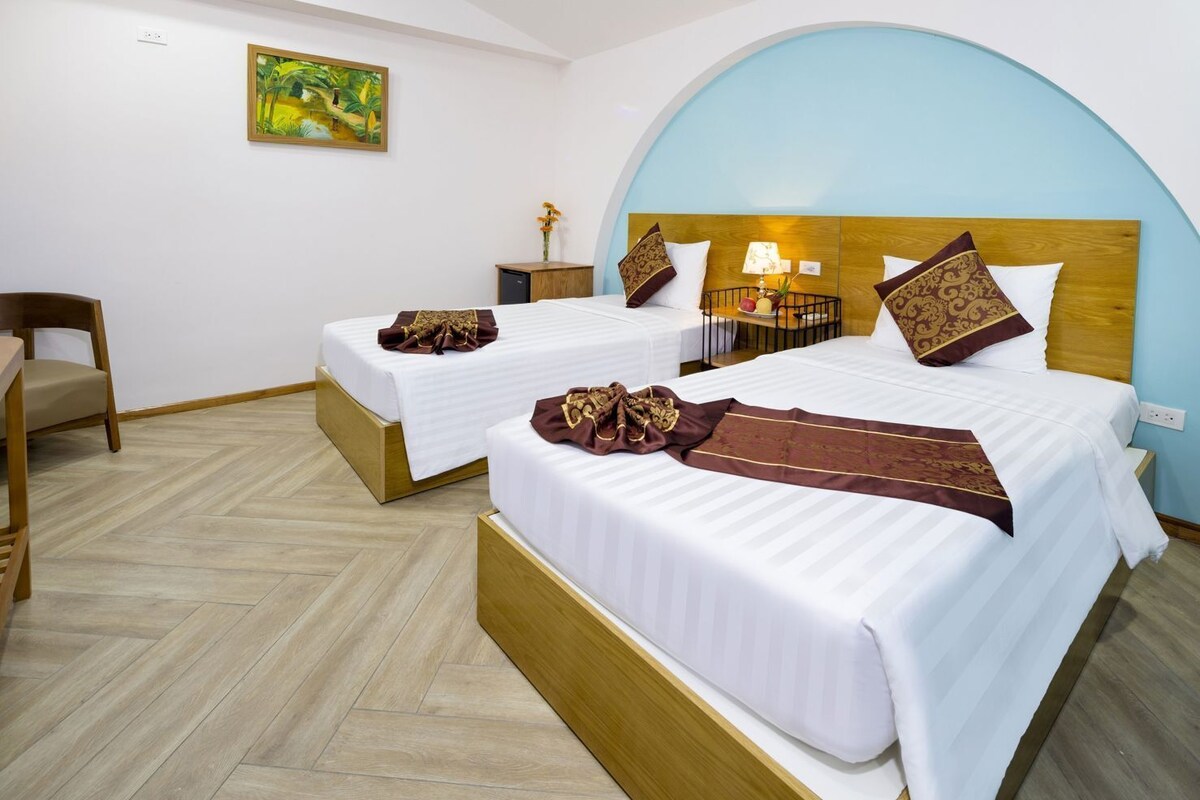 A hotel room with two beds, a wooden headboard, a small table with a lamp, and a painting on the wall.