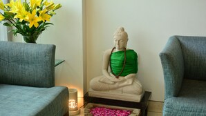 Une statue de Bouddha en position méditative, un vase de fleurs jaunes et deux fauteuils gris dans une pièce dont le sol est recouvert de carreaux.