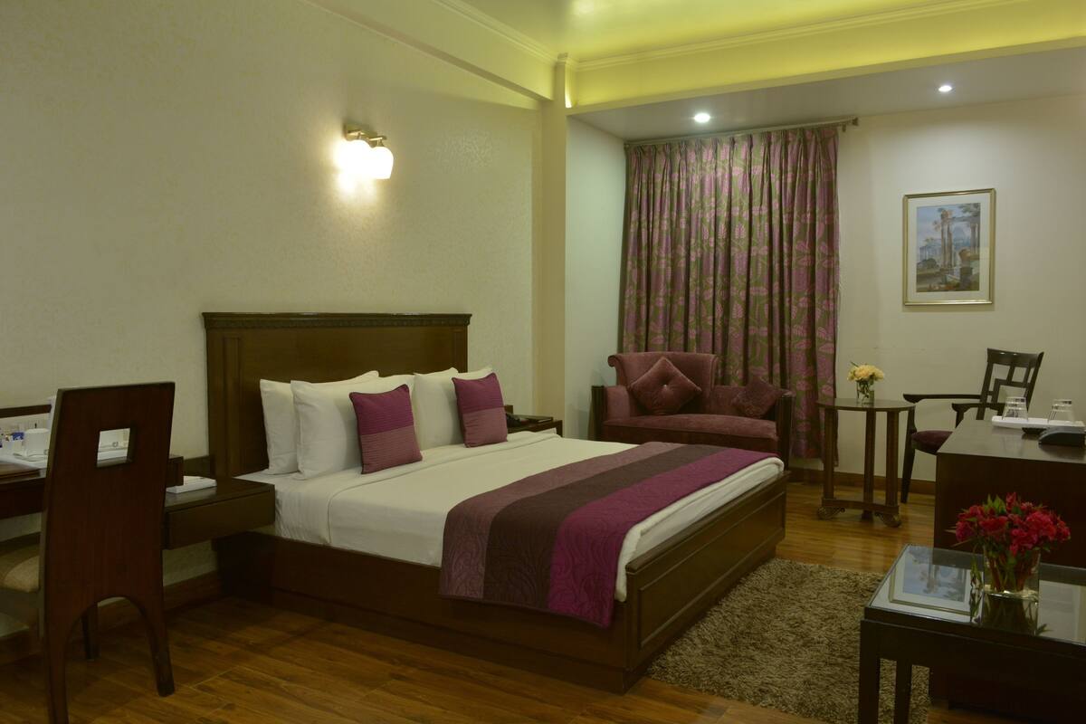 Quarto de hotel com uma cama grande, uma escrivaninha, uma cadeira e uma mesa pequena com um vaso de flores.