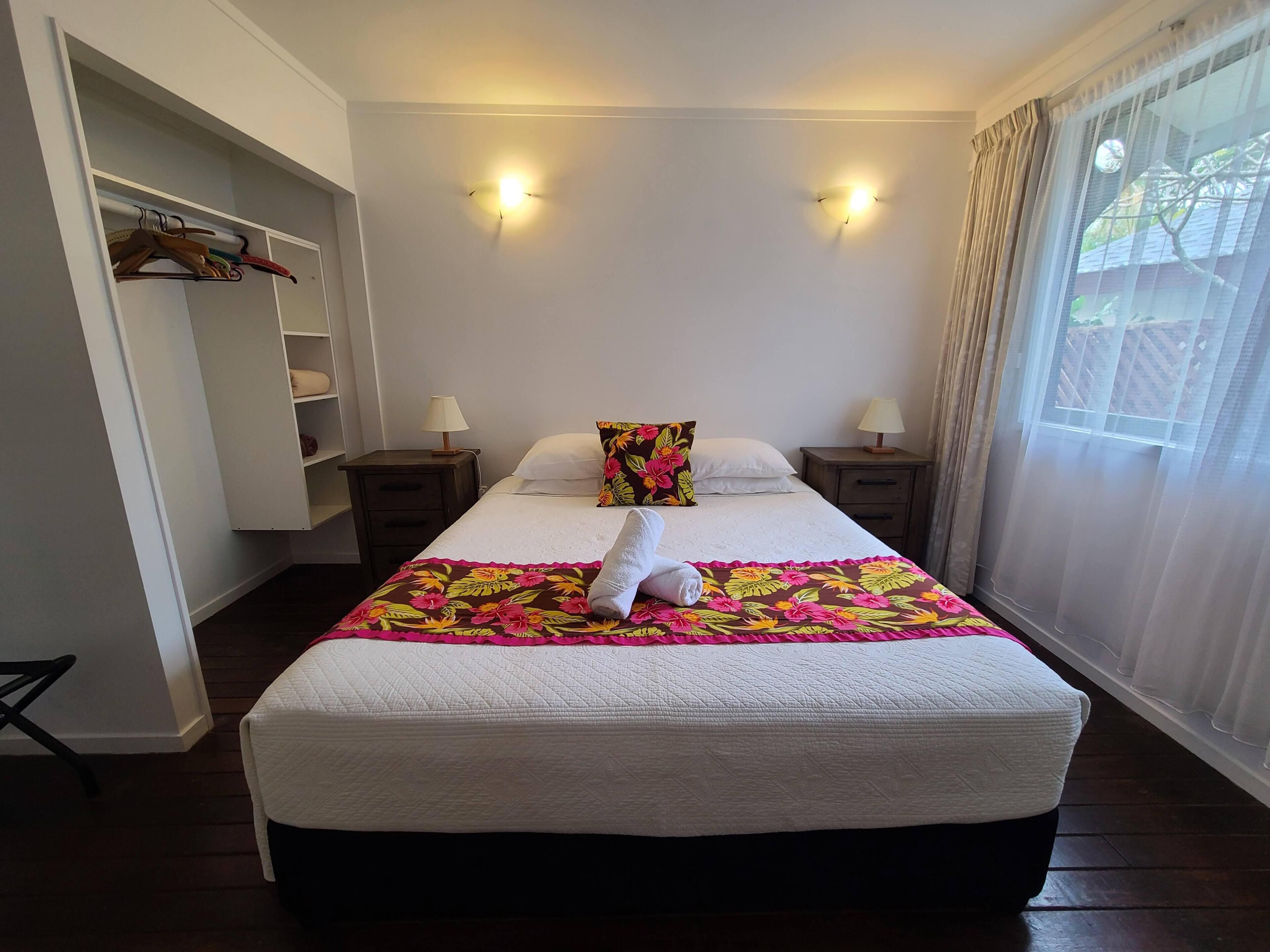 Een opgemaakt bed met een bloemenbedekking, twee bedlampjes en een raam met dunne gordijnen.