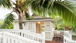 A small house with a wooden deck, a white railing, and a palm tree. - The Cooks Oasis (Rarotonga)