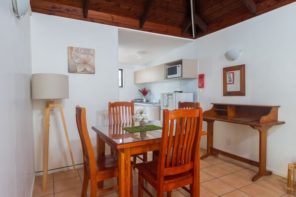 A dining area with a wooden table and chairs, a lamp, and a kitchen in the background. - The Cooks Oasis (Rarotonga)