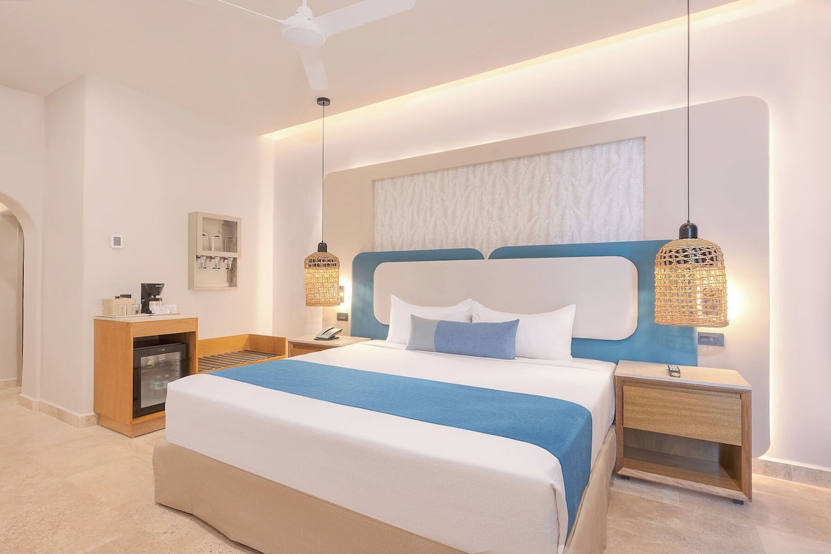 A modern hotel room with a large bed, bedside tables, a minibar, and a ceiling fan.