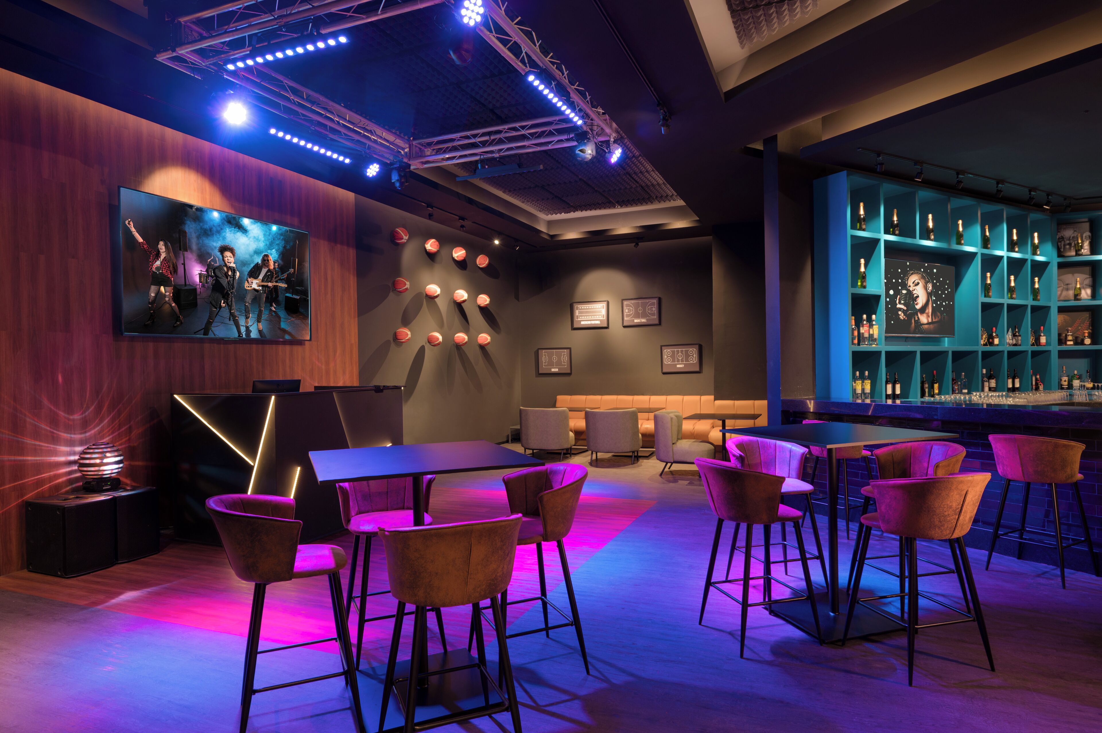 A modern bar area with a large screen displaying a band, bar stools, a counter with bottles, and ambient lighting.