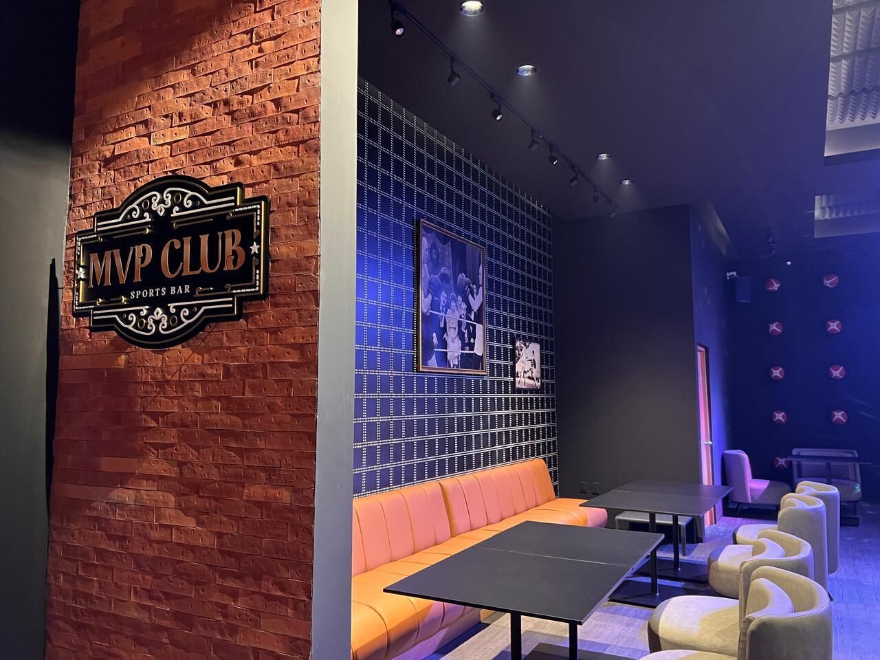 A sports bar with a brick wall, a sign reading 'MVP CLUB SPORTS BAR,' a blue wall with framed pictures, a sofa, and a table with chairs.