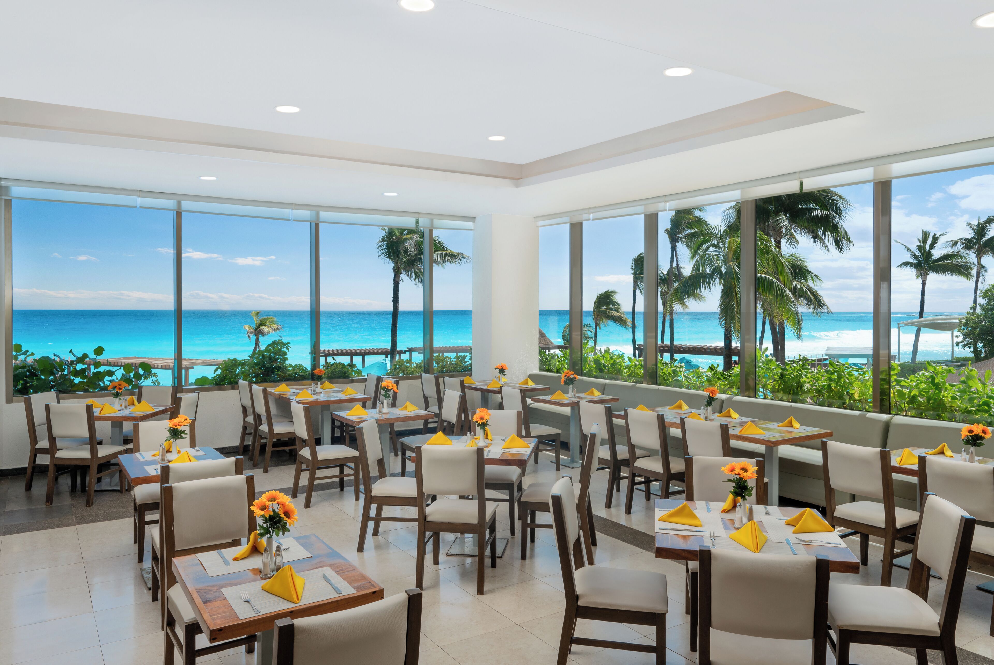 Un restaurant avec des tables et des chaises disposées pour accueillir les clients, de grandes fenêtres offrant une vue sur l’océan et les palmiers.