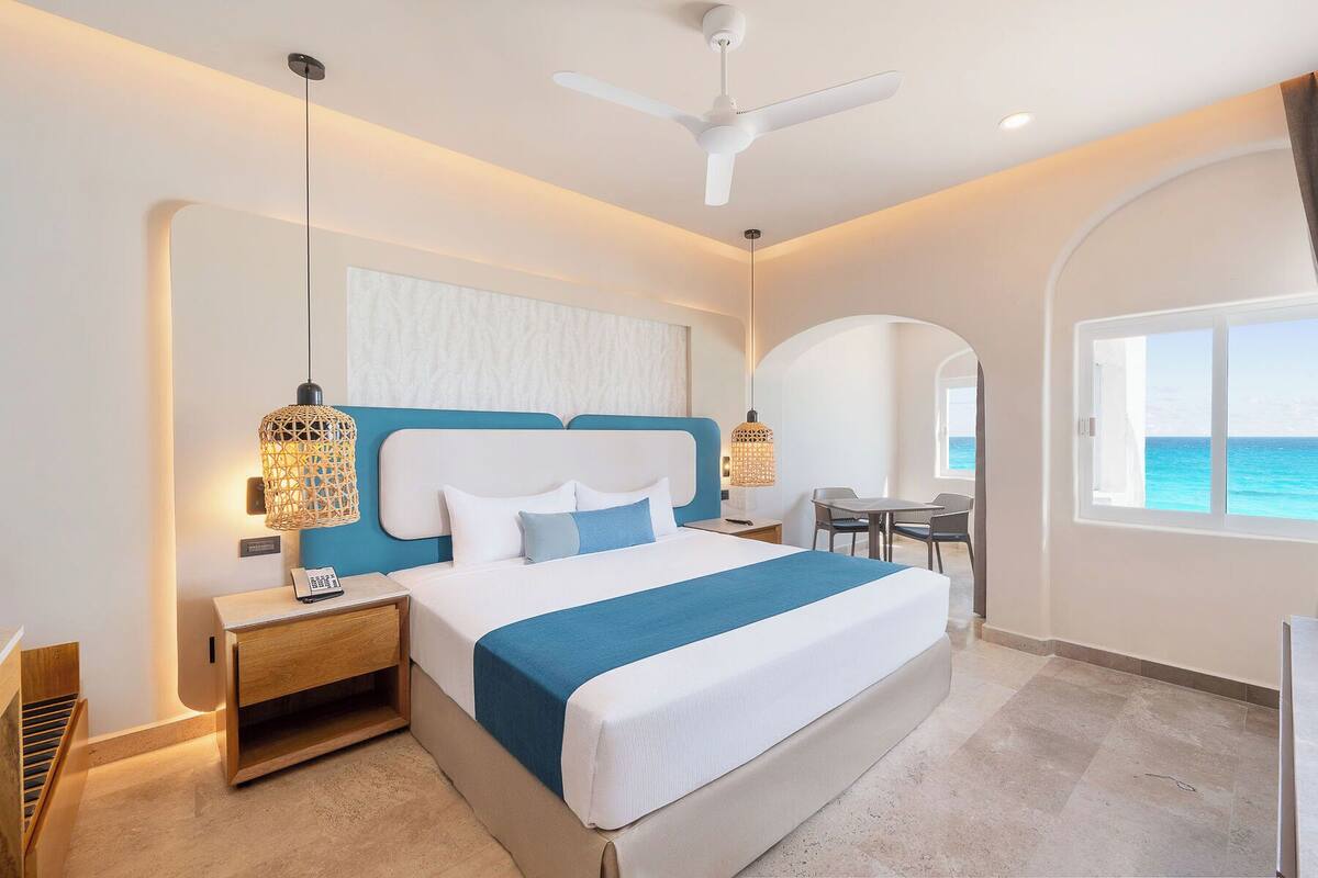 A modern hotel room with a large bed, two bedside tables, a ceiling fan, and a view of the ocean.