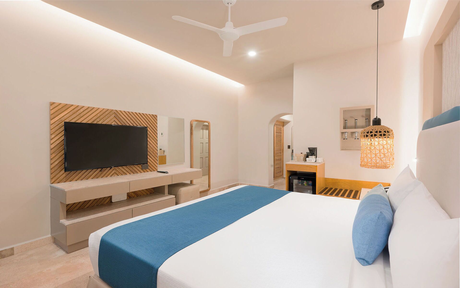 A modern hotel room with a bed, TV, desk, and kitchenette.