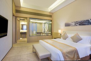 Een hotelkamer met een groot bed, een bankje, een televisie, een badkamer met een spiegel en een kunstwerk aan de muur.