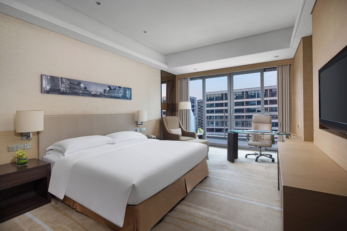 A hotel room with a large bed, a desk, a chair, a TV, and a window with a city view.