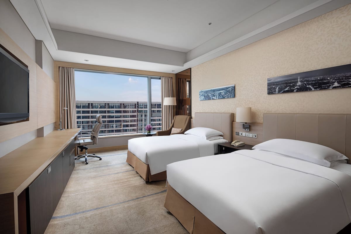 Une chambre d’hôtel avec deux lits, un bureau, une chaise, une télévision et une grande fenêtre donnant sur la ville.
