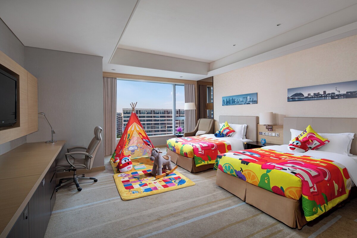 A hotel room with two beds, a desk, a chair, a desk lamp, a window with a city view, and a small play area with a tent and toys.