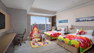 A hotel room with two beds, a desk, a chair, a desk lamp, a window with a city view, and a small play area with a tent and toys.
