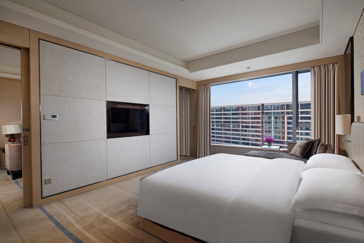 A modern hotel room with a large bed, a flat-screen TV, a view of the cityscape, and a window with curtains.