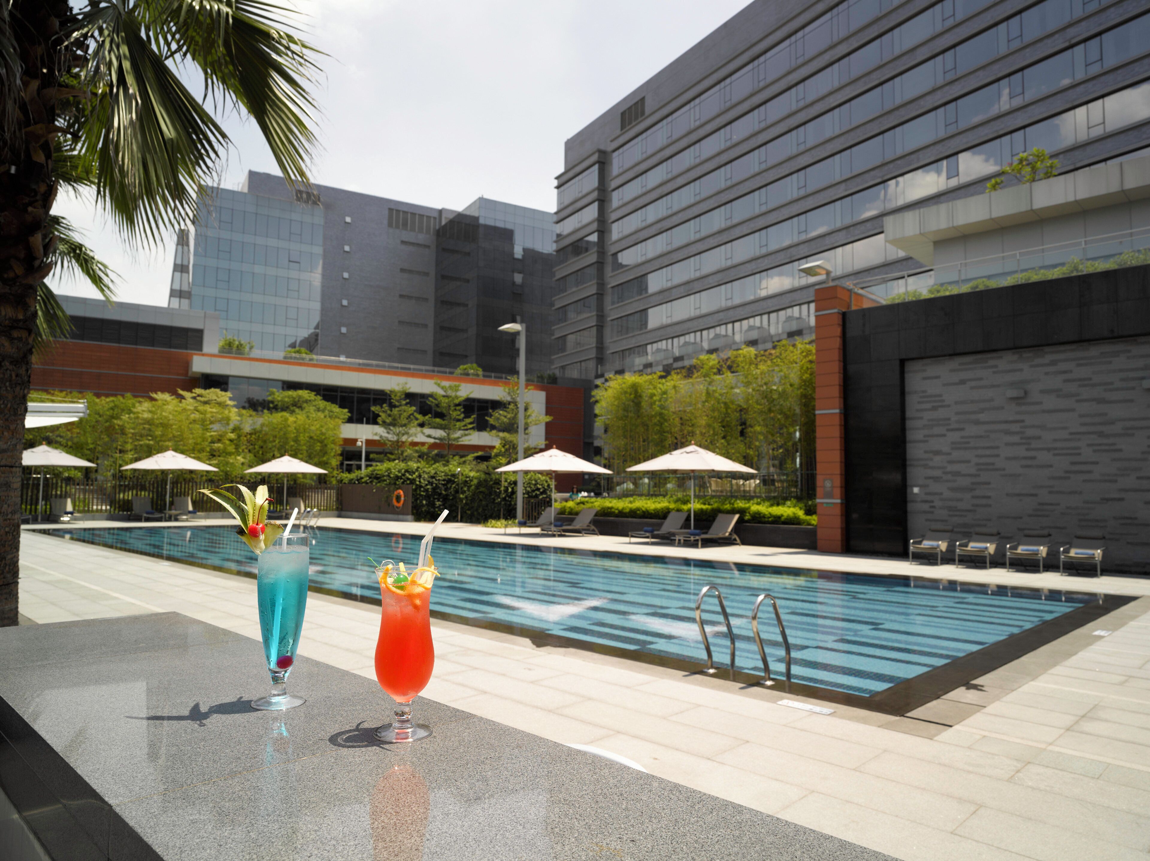 Zona junto a la piscina con dos bebidas sobre una mesa, sillas de descanso y un edificio moderno al fondo.