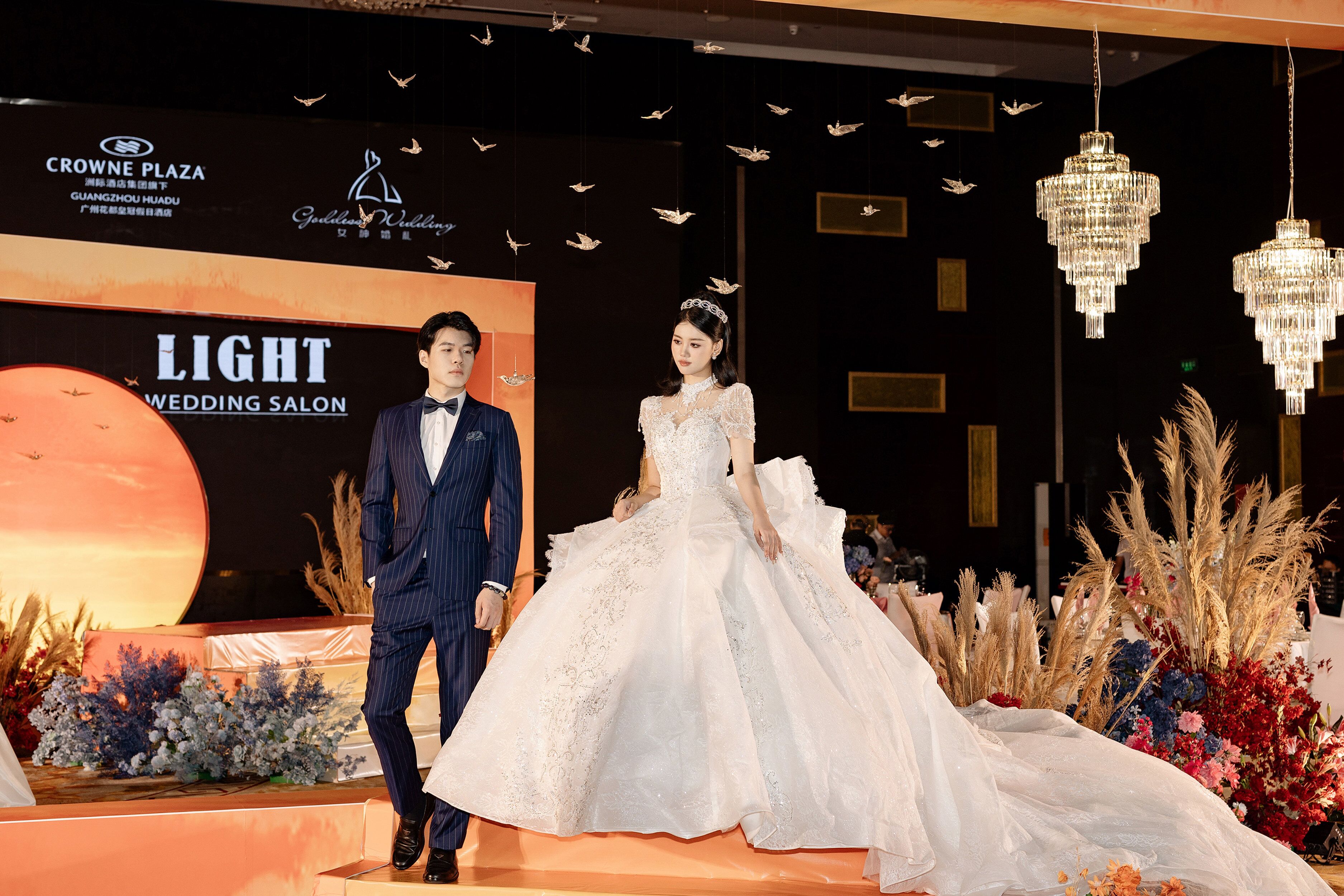 A wedding scene with a couple on a stage, a large illuminated backdrop, and hanging chandeliers.