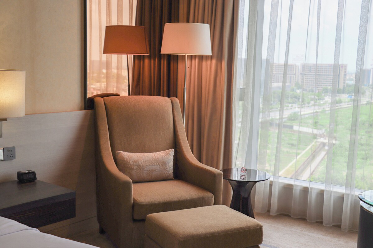A hotel room with a large window, a chair, a small table, and a lamp.
