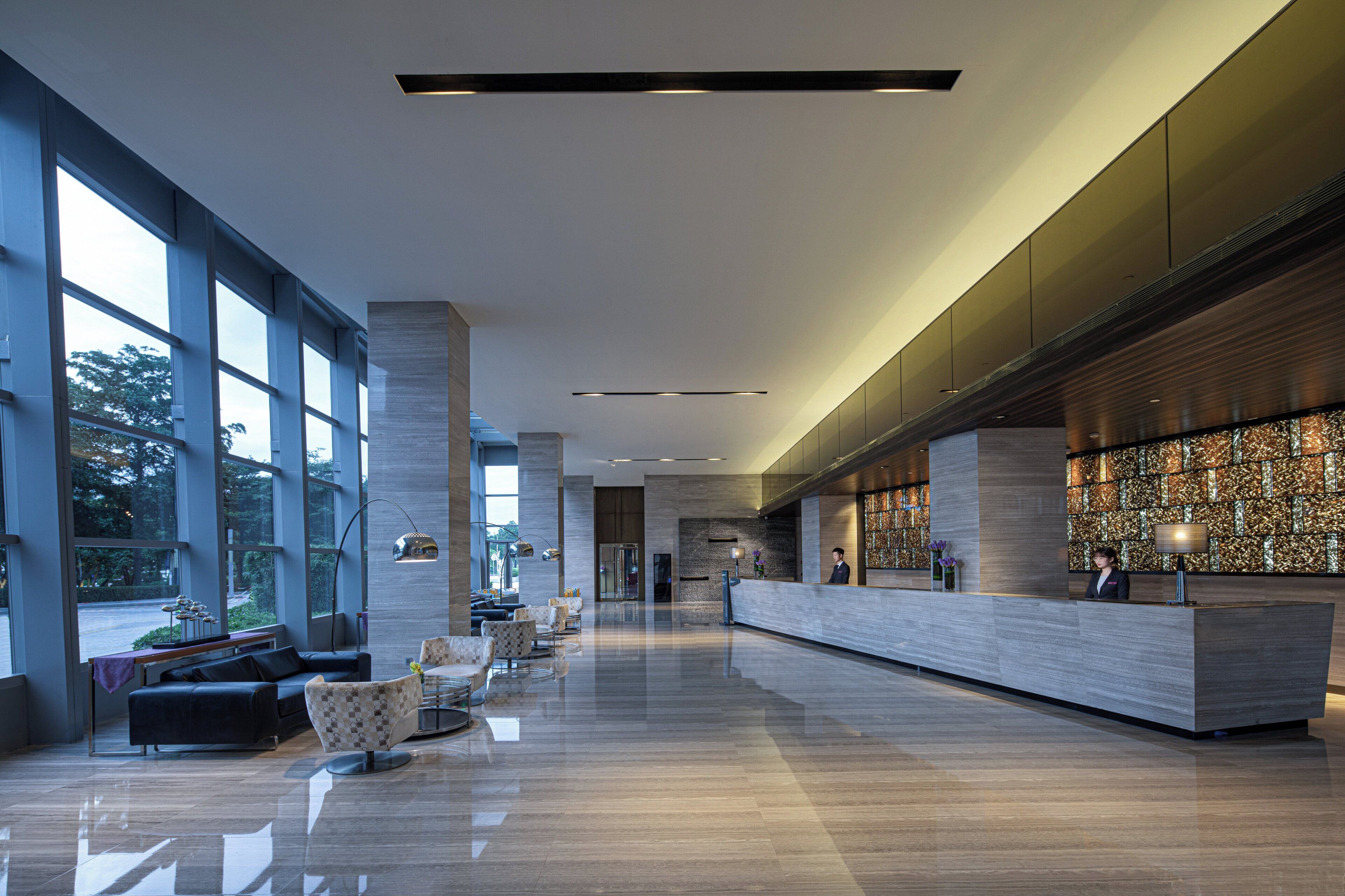 A modern hotel lobby with a reception desk, seating area, and large windows.