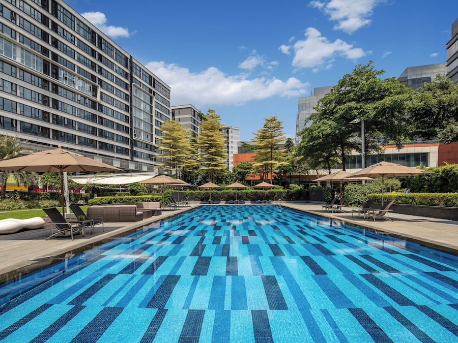 A swimming pool with a blue tiled pattern, surrounded by lounge chairs and umbrellas, with modern buildings in the background.
