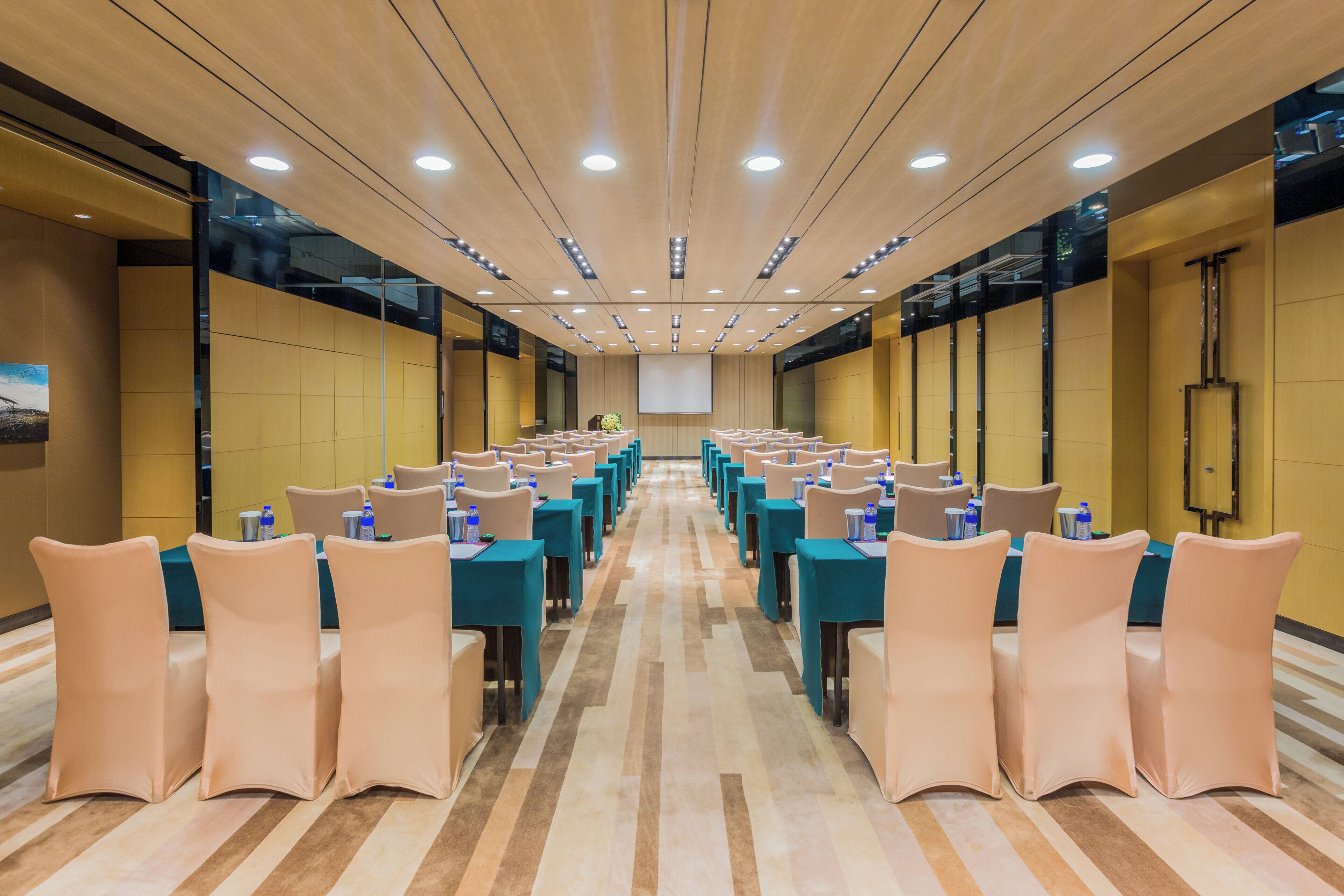 Une salle de conférence avec des rangées de tables et de chaises, équipée de bouteilles d’eau et dotée d’un grand écran à l’avant.
