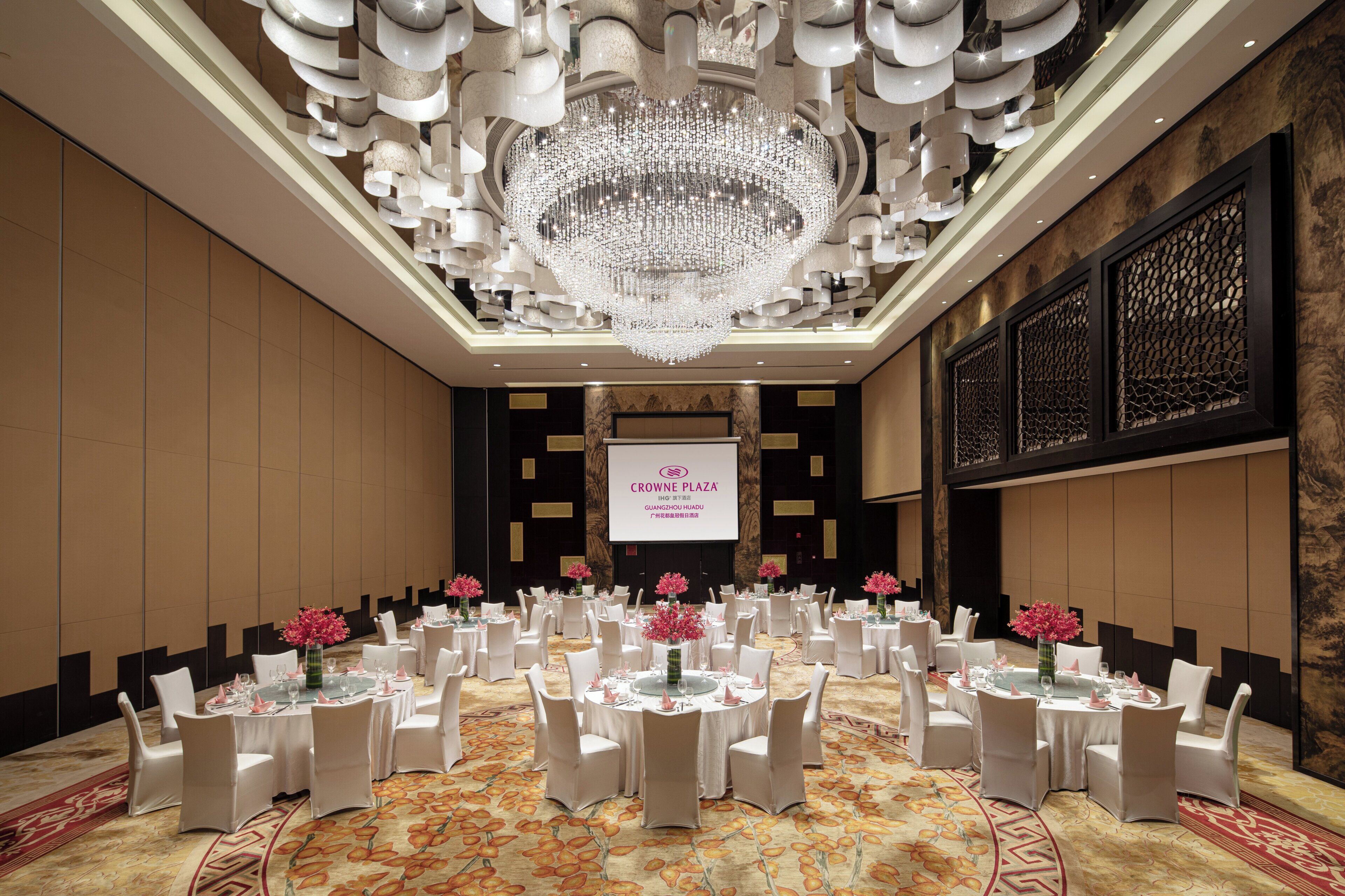 A banquet hall with round tables, white chairs, and a large chandelier.