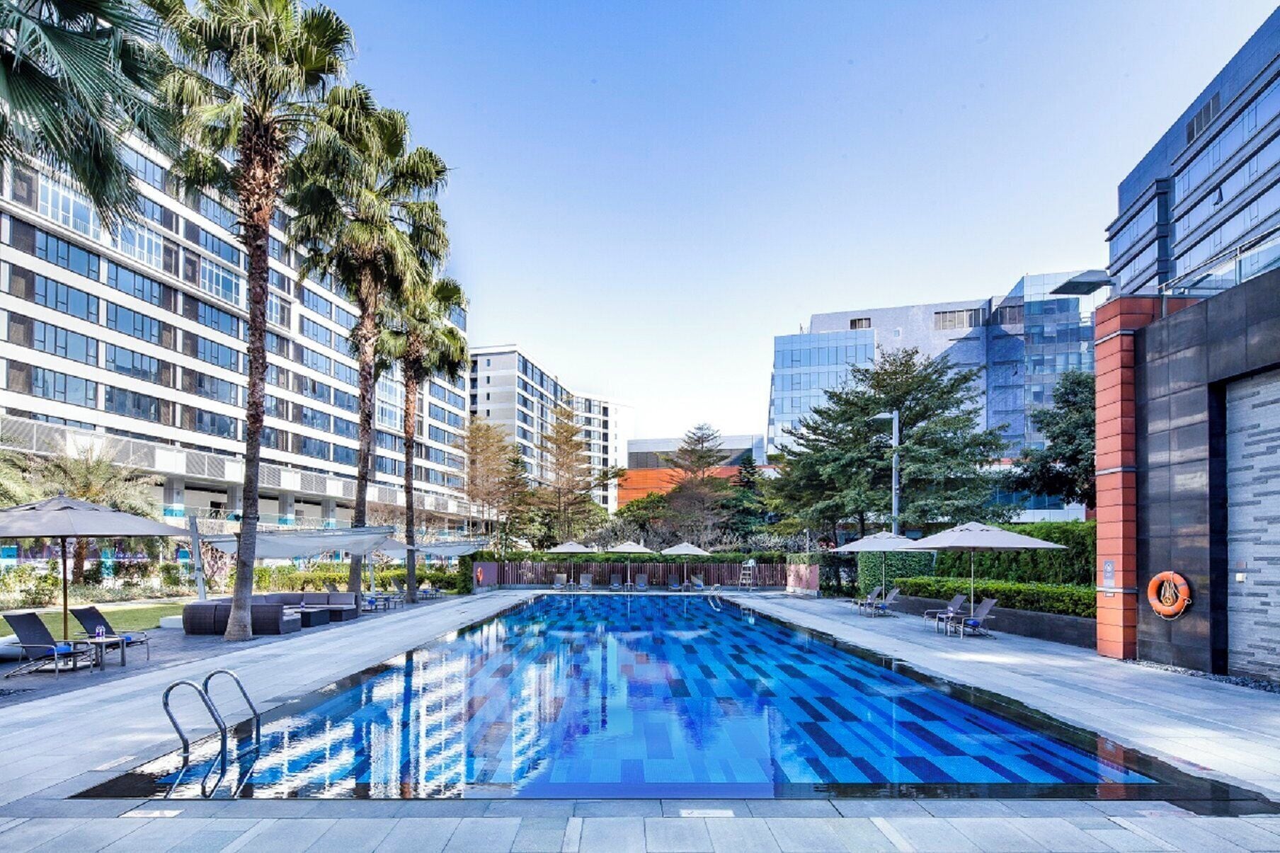 Una piscina con sillas de descanso y sombrillas, rodeada de edificios modernos y palmeras.